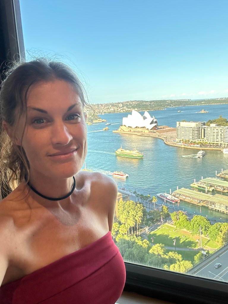 A woman smiling in front of a window with a view of the Sydney Opera House and the harbor.
