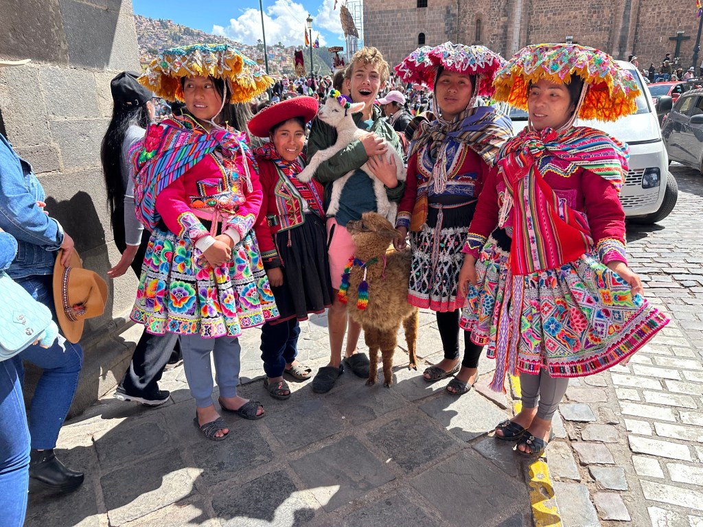 A group of local women in traditional colorful Peruvian attire, including skirts and hats, pose for a photo with a young boy holding a small llama in Cusco, Peru.