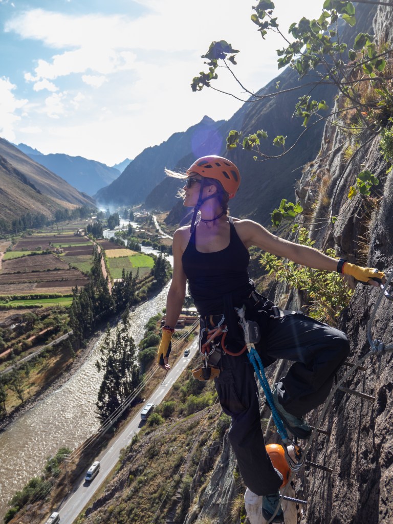 A person climbing a via ferrata on a rocky cliff in the Andes mountains, wearing a helmet and climbing gear, with a view of a river and lush farmland below.