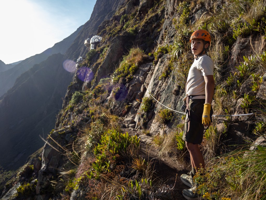 A climber with an orange helmet and gloves stands on a rocky path with lush vegetation, overlooking the Sacred Valley in Peru, with capsule accommodations visible on the cliffside.