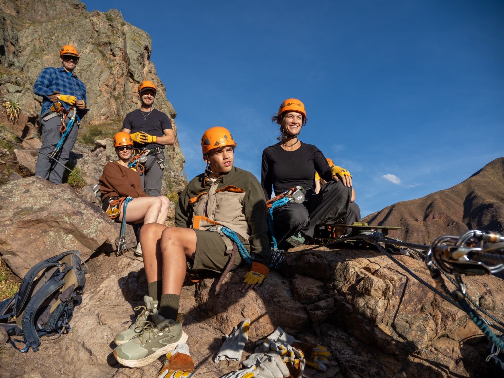 A group of people wearing climbing harnesses and helmets, resting on a rocky cliff in the Sacred Valley of Peru, with mountainous terrain and blue skies in the background.