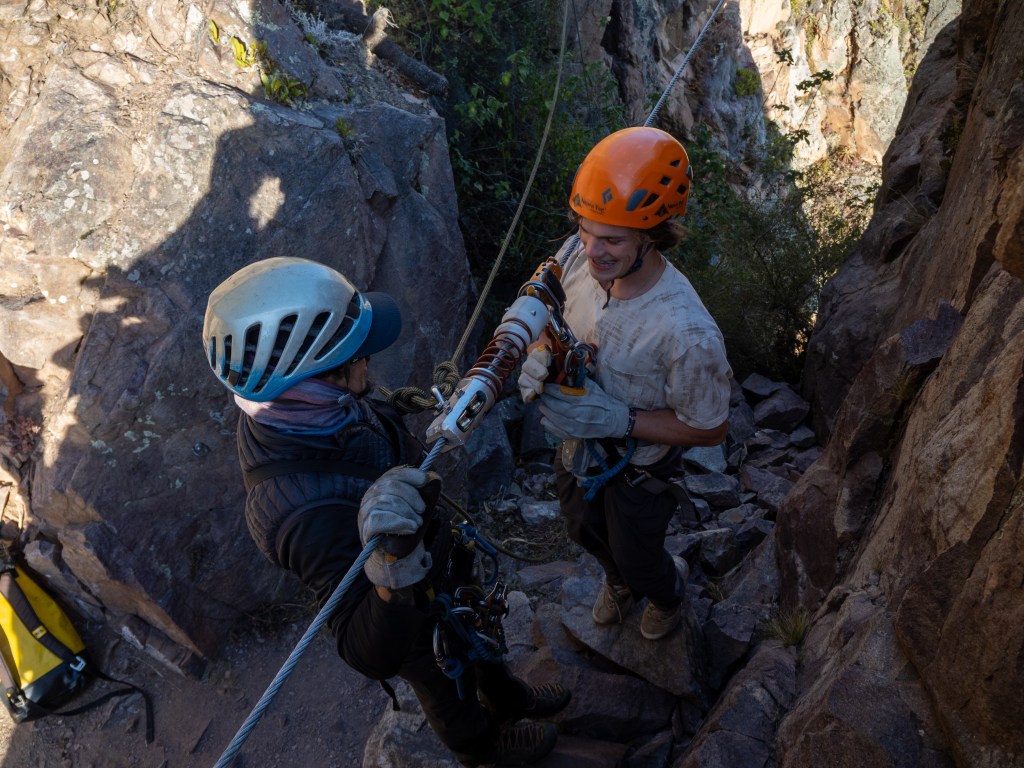 Two climbers are seen preparing to ascend a rocky cliff, one wearing a helmet and harness while the other is holding climbing equipment. Sunlight filters through the canyon, illuminating the scene.