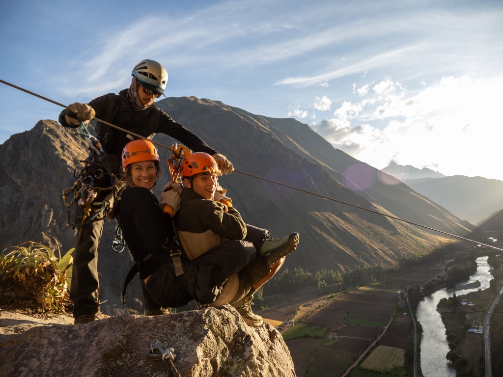 Two people in harnesses preparing for a zipline descent, with a mountain backdrop and river below in the Sacred Valley, Peru.