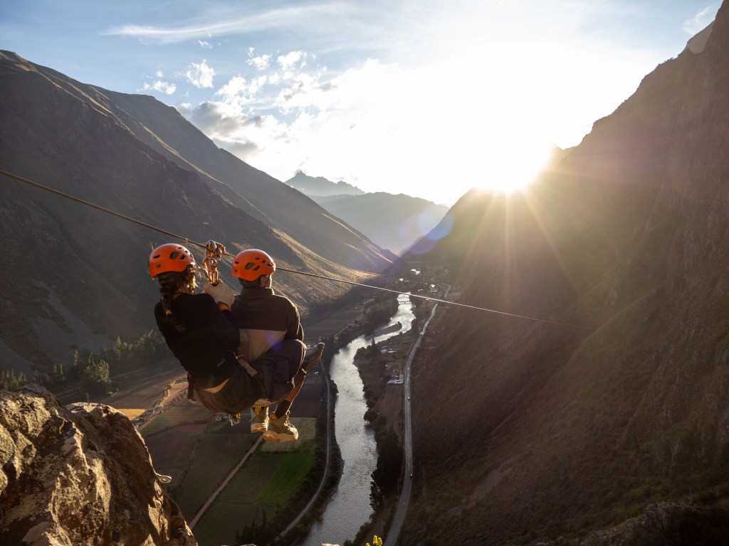 Two people ziplining across a scenic valley with mountains and a river below, bathed in golden sunlight.