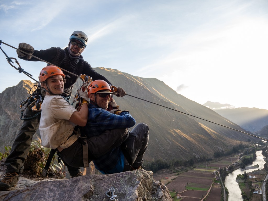 Three individuals enjoying a ziplining experience against a backdrop of mountainous terrain and a valley with a river.