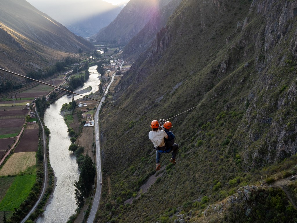 Two people ziplining over the Sacred Valley in Peru, with mountains and a winding river visible below.