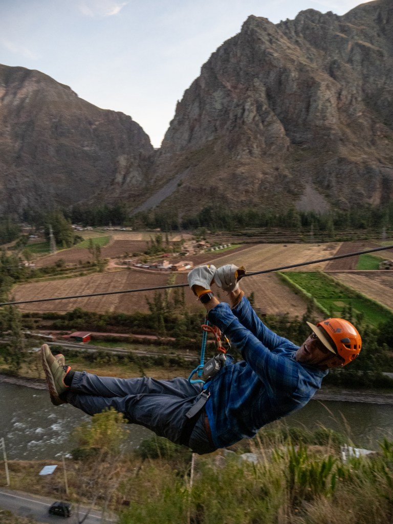 A man ziplining over the Sacred Valley in Peru, with mountains and agricultural land visible in the background.