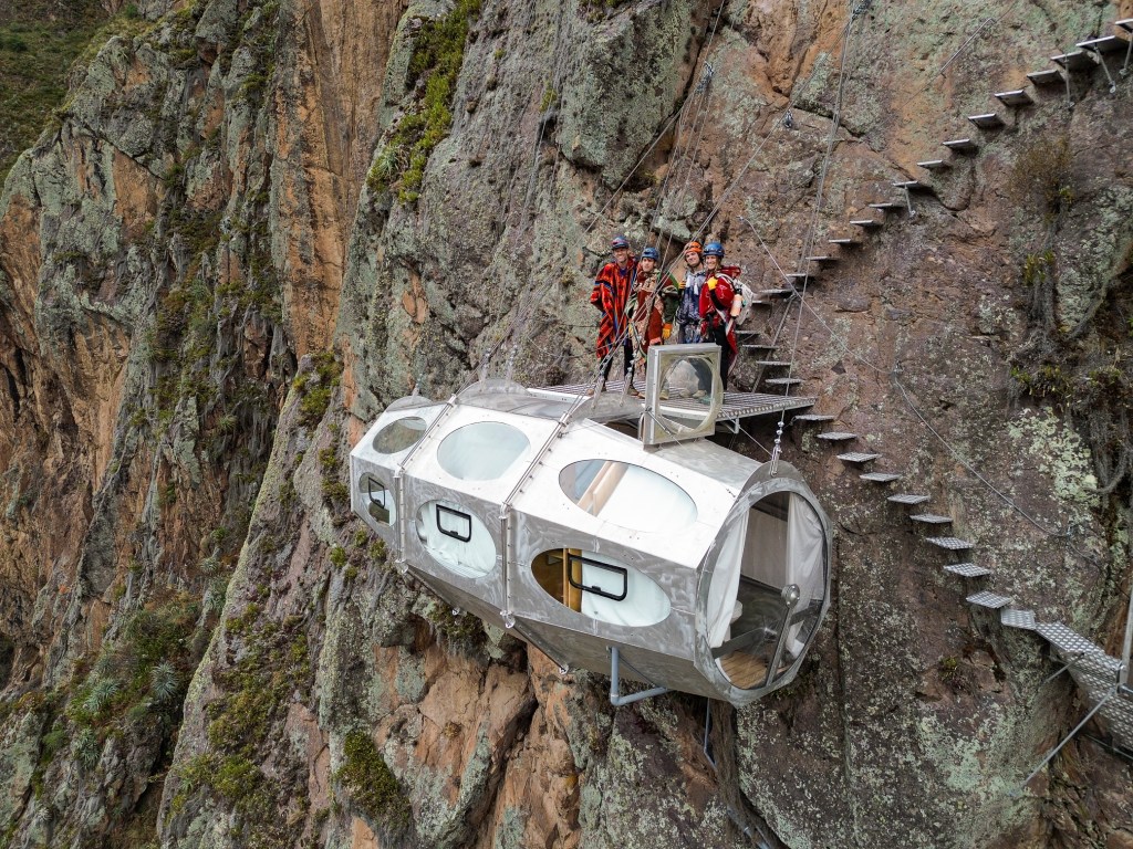 A group of four climbers standing on a transparent, pod-like accommodation suspended on a cliffside in the Sacred Valley, Peru, with rocky terrain and a staircase leading up the mountain.