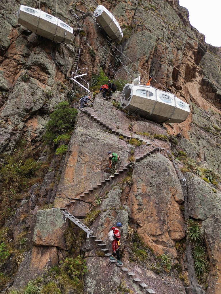 Climbers ascending rugged steps on a cliffside, with glass capsules suspended above, near Skylodge and Starlodge in the Sacred Valley, Peru.