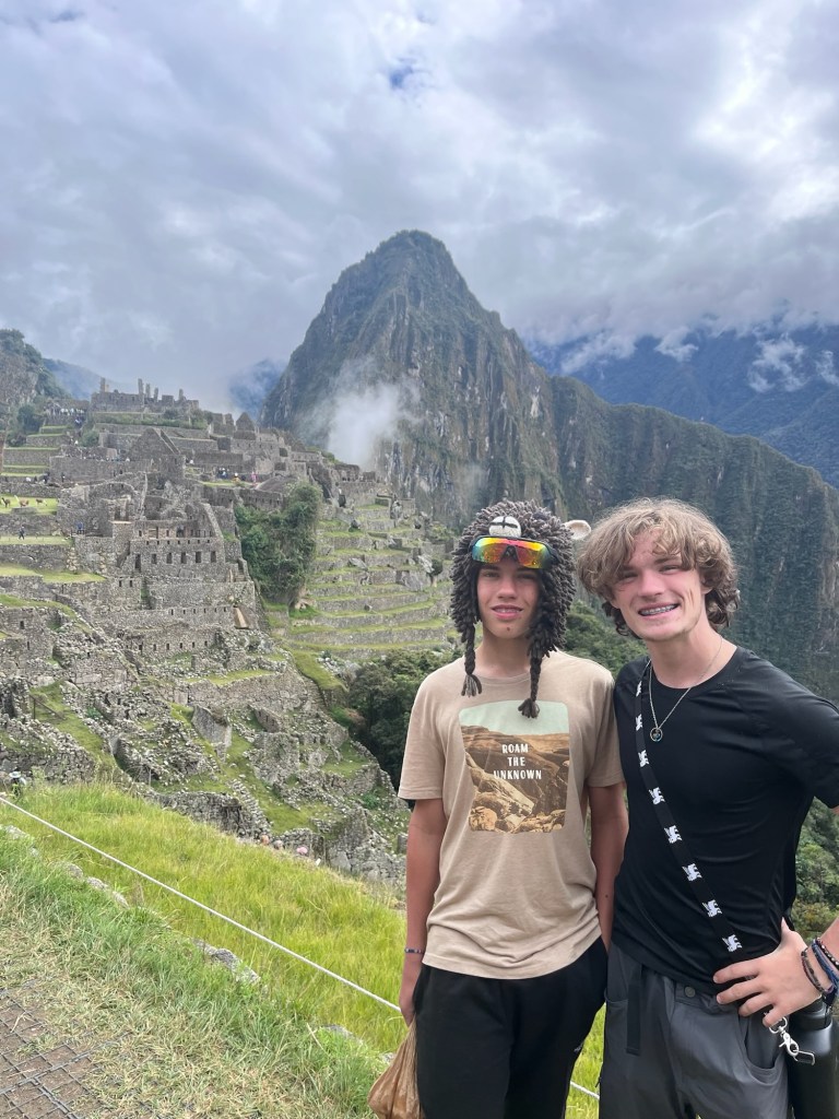 Two young travelers pose in front of the Machu Picchu ruins, with dramatic mountains and cloudy skies in the backdrop.