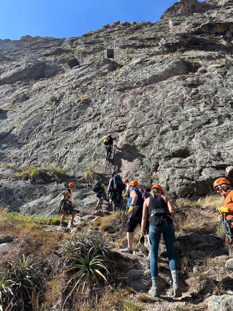 Group of climbers wearing helmets and harnesses ascending a rocky cliff in Sacred Valley, Peru, with supportive climbing gear visible.