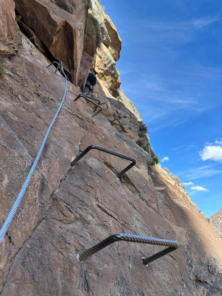 Close-up view of a rocky cliffside with metal handholds installed for climbing, accompanied by a rope secured to the rock.