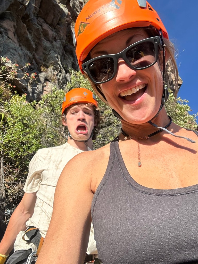 A person wearing an orange climbing helmet and sunglasses smiles for a selfie while a second person in the background makes a funny face, both surrounded by rocky terrain and greenery.
