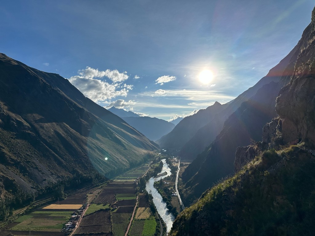 A breathtaking view of the Sacred Valley in Peru, featuring rolling mountains, lush green fields, and a winding river under a bright blue sky with the sun shining.