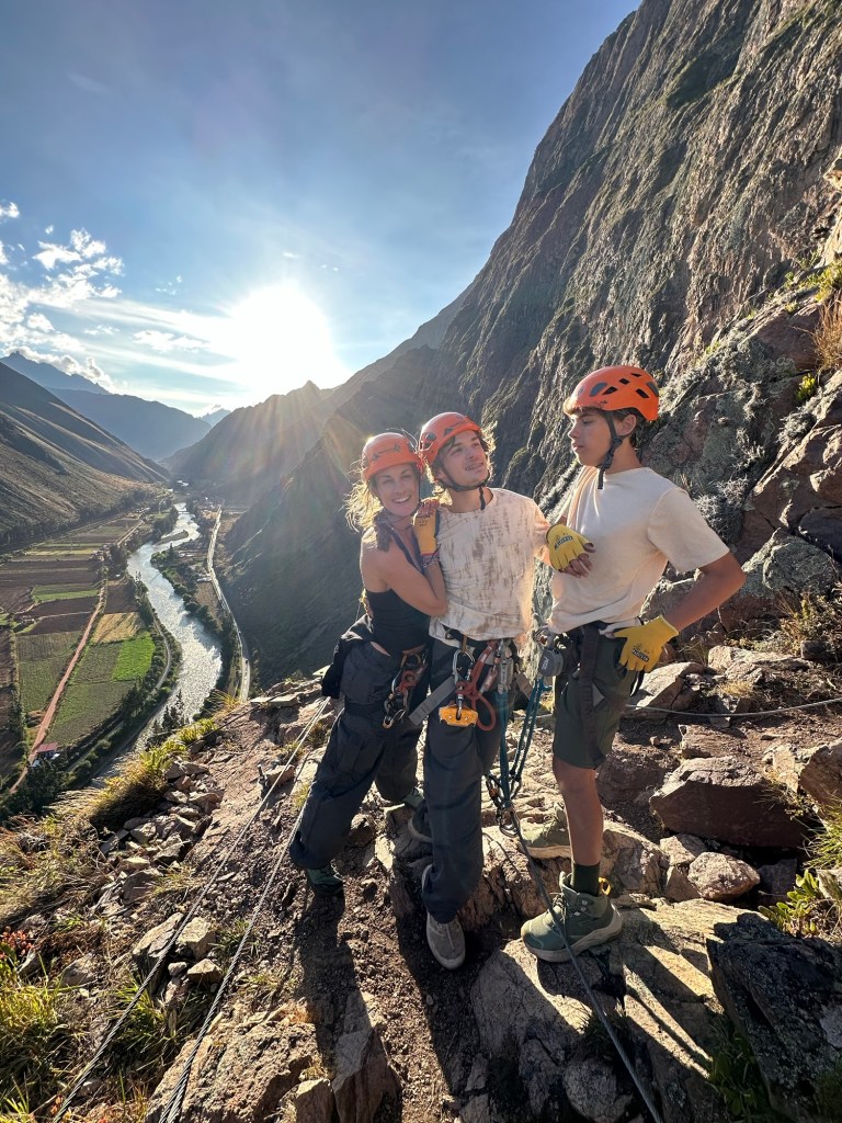 Three climbers posing on a rocky ledge during a Via Ferrata adventure, with the Sacred Valley and a river in the background under a sunny sky.