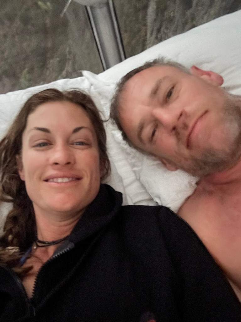 A close-up selfie of a man and woman in a cozy, glass capsule, smiling and enjoying their morning together.