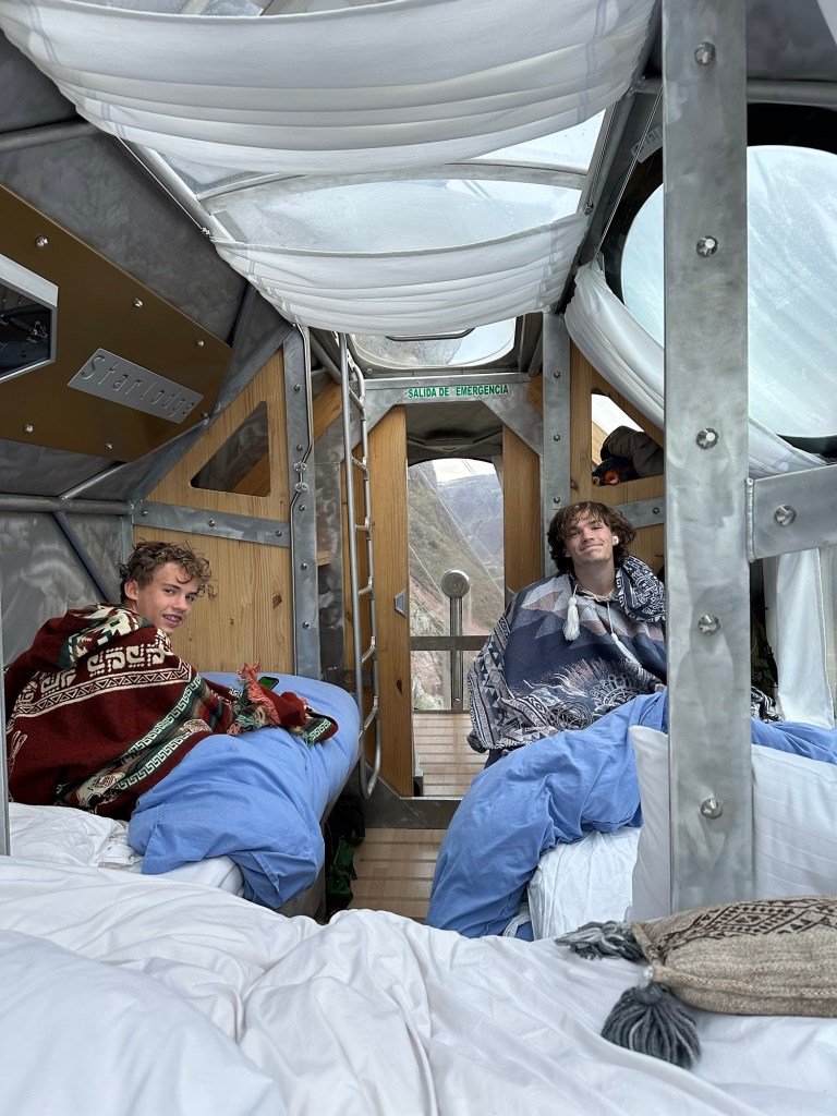 Two young men sitting inside a unique capsule accommodation at Starlodge, wrapped in blankets, with a view of the Sacred Valley visible through large windows.