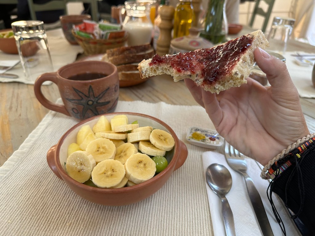A hand holding a slice of bread topped with jam, paired with a bowl of sliced bananas and green grapes, and a cup of coffee on a dining table.