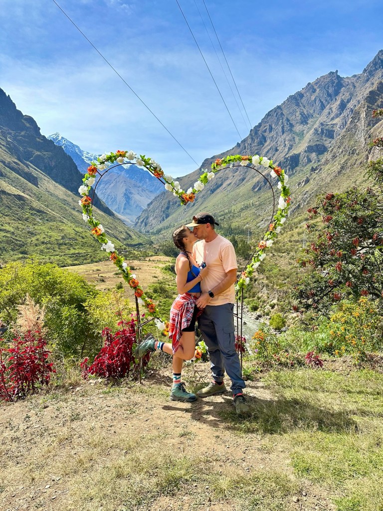A couple embraces in front of a heart-shaped floral arch surrounded by mountains and greenery in Peru.
