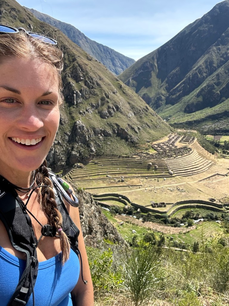 Person smiling on a hiking trail with a scenic mountainous background.