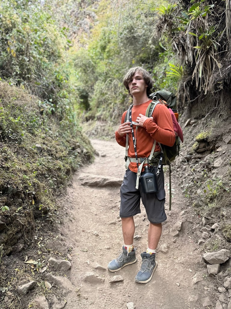 A young hiker stands on a rocky path in a lush, green landscape, wearing a red long-sleeve shirt and carrying a backpack.