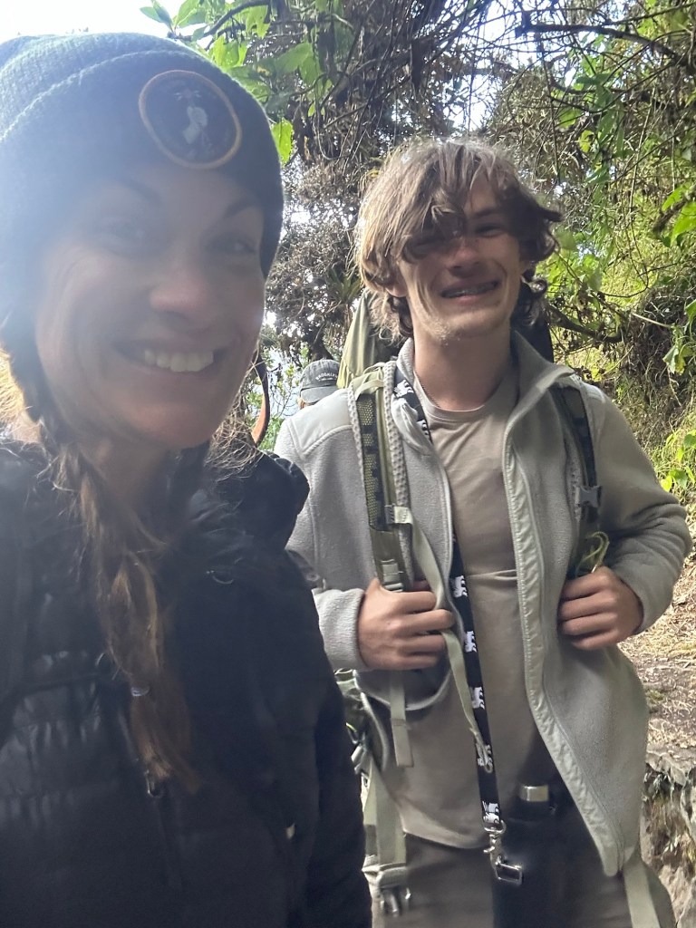 A smiling woman and a young man posing for a selfie on a hiking trail, surrounded by lush greenery.