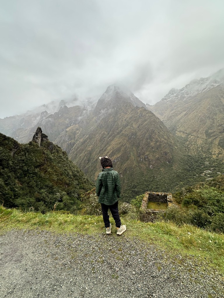 A person standing on a mountainous trail overlooking lush green hills and mountains under a cloudy sky in Peru.