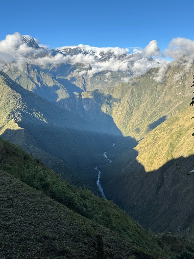 A breathtaking view of a deep valley in Peru, surrounded by towering mountains with snow-capped peaks and fluffy clouds in a clear blue sky.