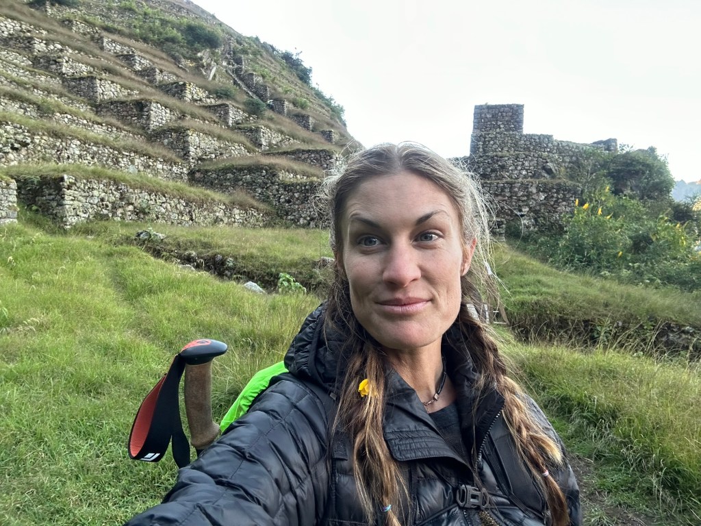 A woman taking a selfie in a mountainous area with terraced stone structures in the background, surrounded by greenery.