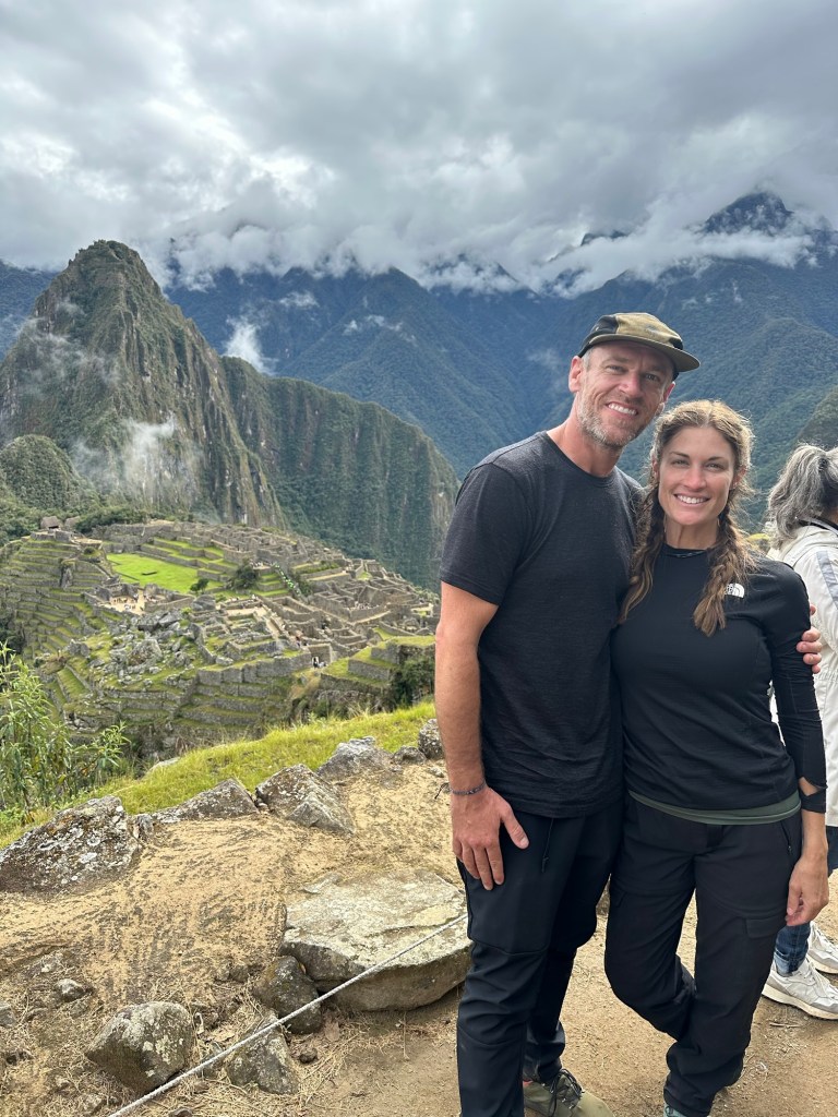 A couple standing at Machu Picchu with lush mountains and ancient ruins in the background.