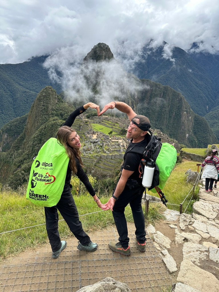 A couple at Machu Picchu, forming a heart shape with their hands while wearing backpacks, with the ancient ruins in the background and lush green mountains shrouded in clouds.