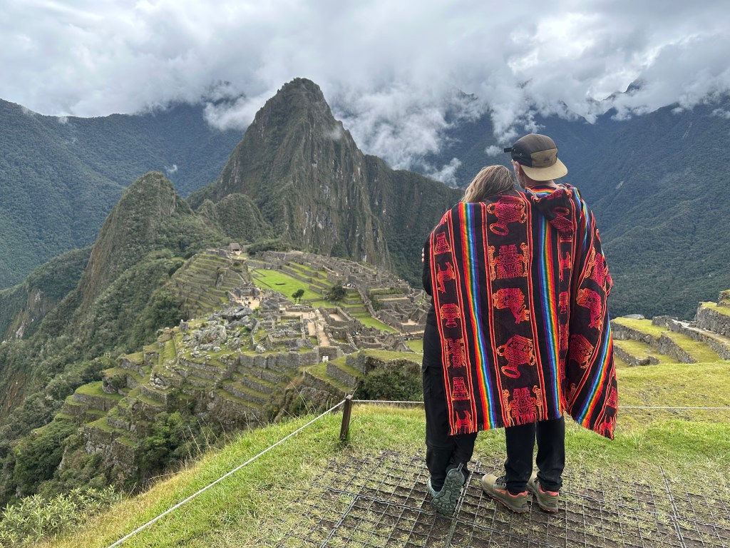 A view of Machu Picchu, showcasing the ancient ruins surrounded by lush green mountains under a cloudy sky, with two individuals wearing colorful ponchos standing in the foreground.