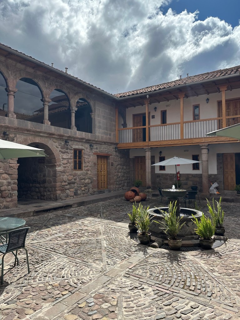 A scenic view of a courtyard in Cusco, Peru featuring rustic stone walls, wooden balconies, and potted plants surrounding a central fountain, under a partly cloudy sky.