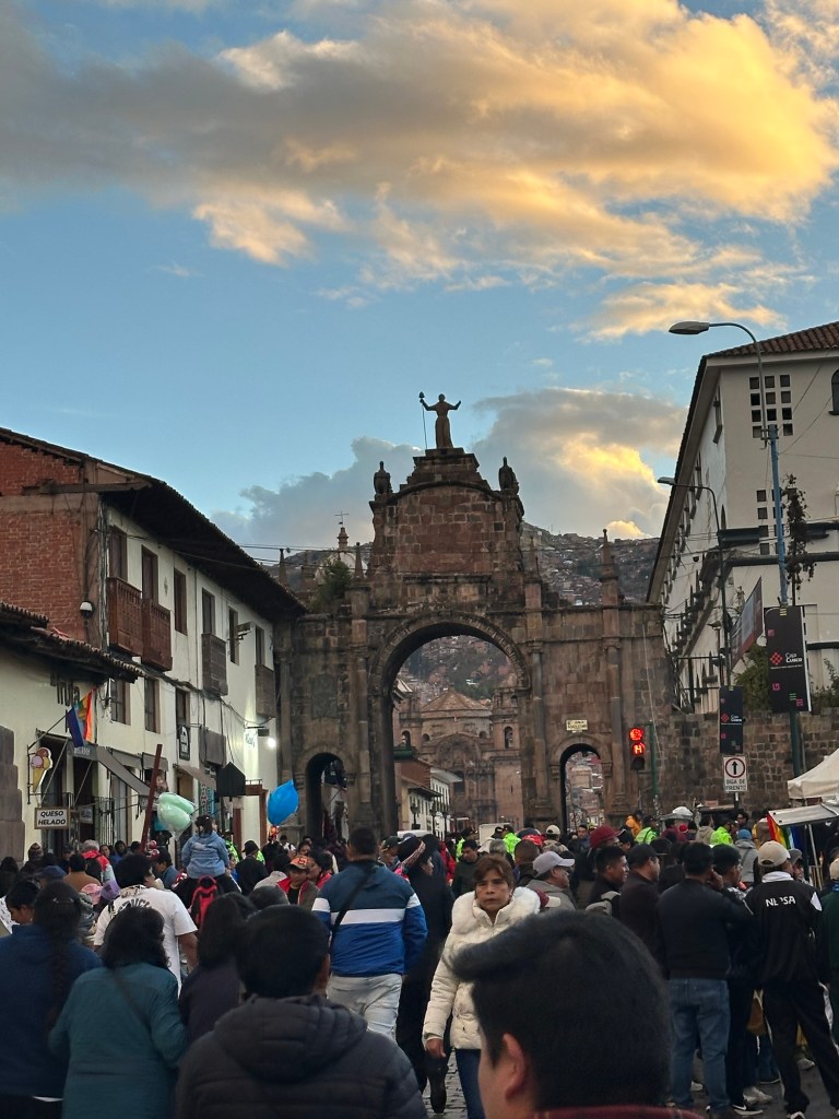 A bustling street scene in Cusco, Peru, showcasing a crowd of people gathered around a historic archway against a backdrop of colorful clouds at sunset.