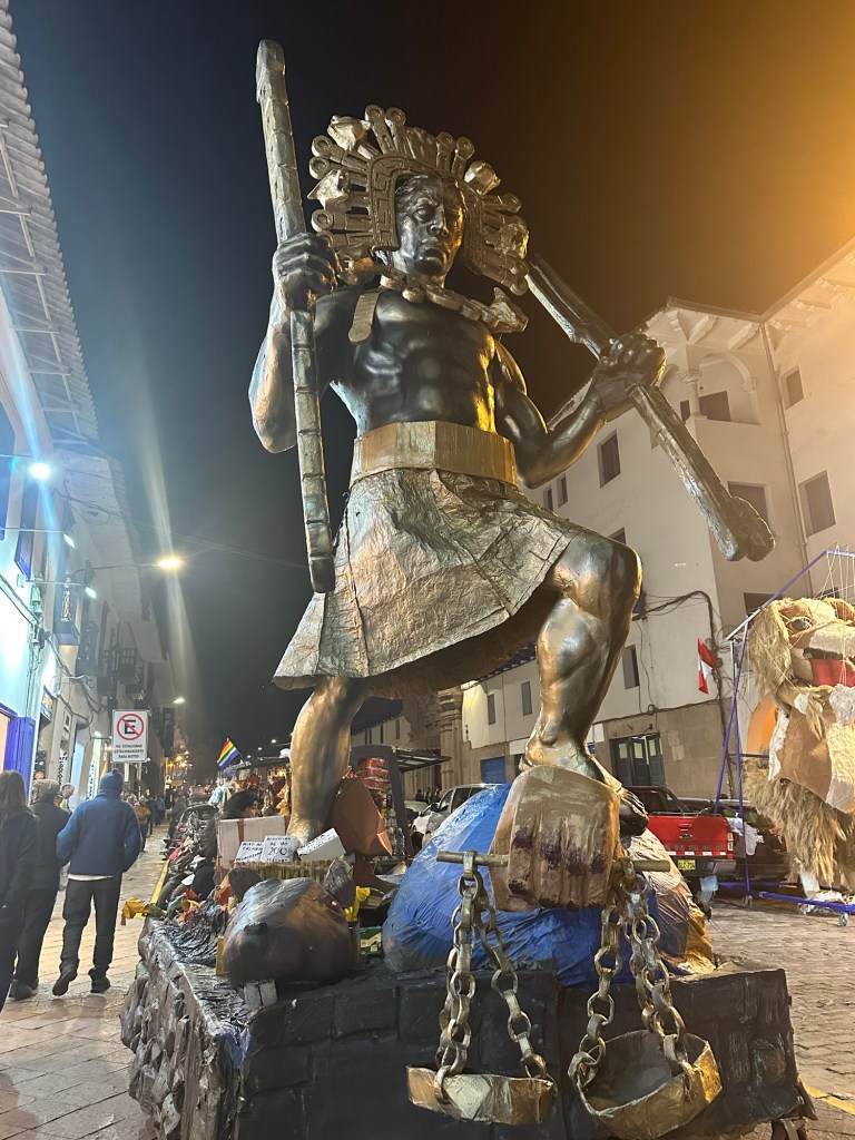 A large statue of an indigenous warrior in traditional attire, holding a spear and standing on a pedestal, illuminated at night with people in the background.