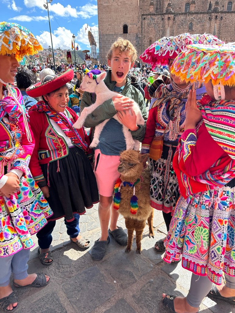 A group of young travelers joyfully interacting with locals in traditional Peruvian attire while holding a fluffy white baby alpaca and a small llama, with a lively crowd and Cusco's architecture in the background.