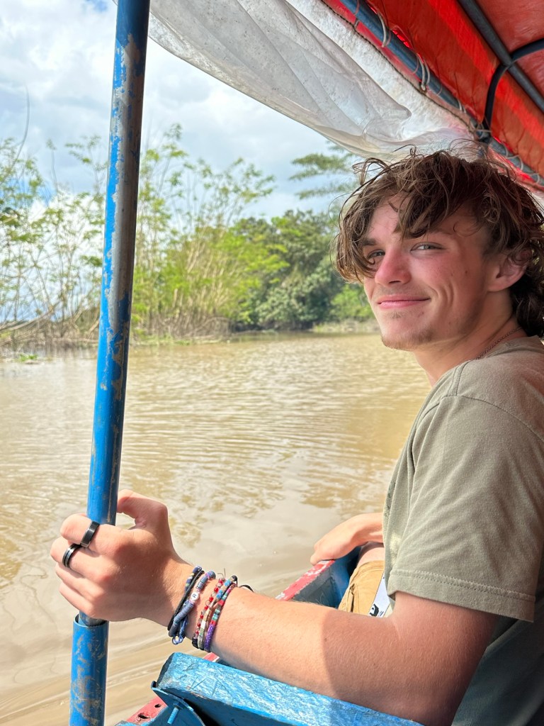 A young person sitting in a boat on a river, smiling and holding onto a blue pole, surrounded by greenery.