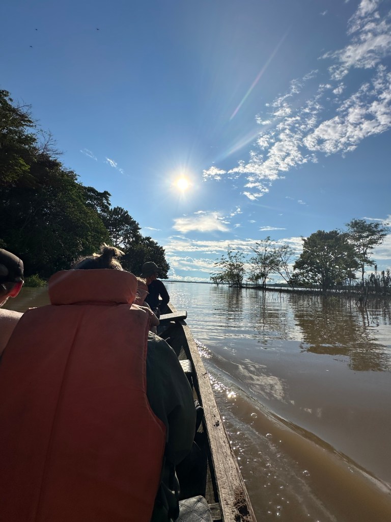 A boat navigates through the Amazon River under a bright sun with clear blue skies and scattered clouds, surrounded by lush greenery and trees on the riverbanks.