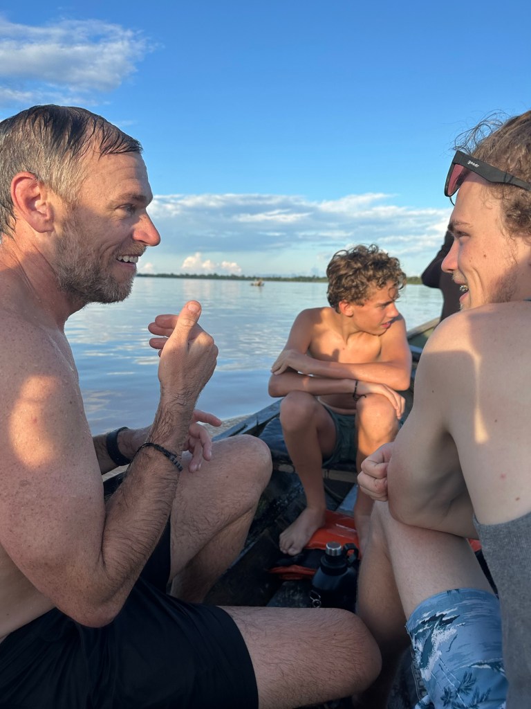 A group of three young boys and an adult, seated in a boat during a sunny day on the Amazon River, engaged in conversation with a scenic blue sky and clouds in the background.