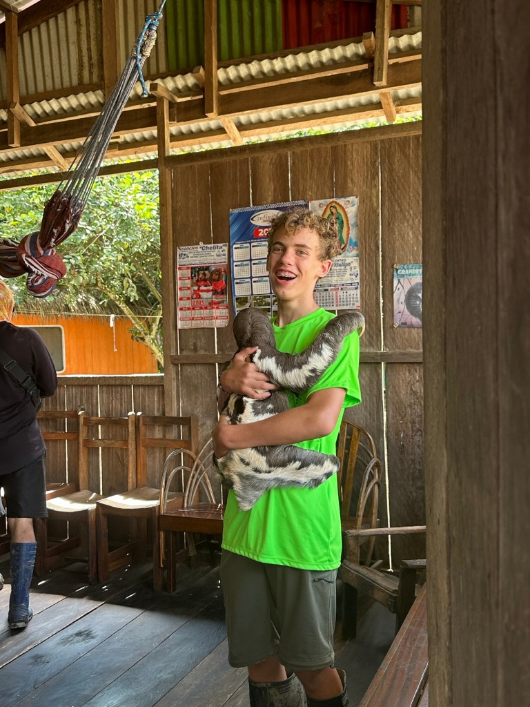 A young boy in a vibrant green shirt happily holds a small sloth in a wooden cabin, surrounded by rustic furniture and walls adorned with posters. The scene captures a moment of joy and connection with wildlife.