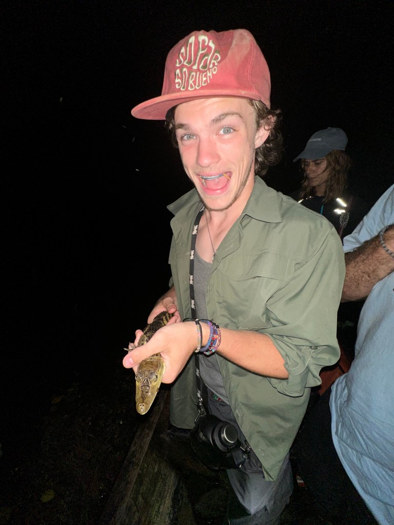 A young man holding a small caiman while smiling, surrounded by others in a nighttime jungle setting.