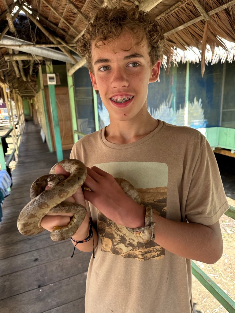 A young boy holding a snake while standing on a wooden walkway in a rustic building surrounded by lush greenery.