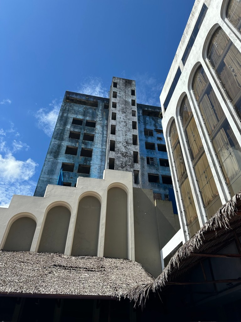 A view of a partially constructed and weathered concrete building against a blue sky, with tropical vegetation in the foreground.
