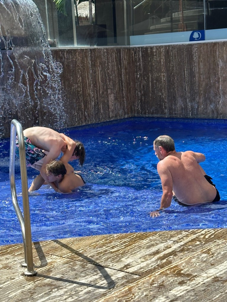 Two young boys play in a swimming pool while an adult supervises nearby, with a waterfall feature in the background.