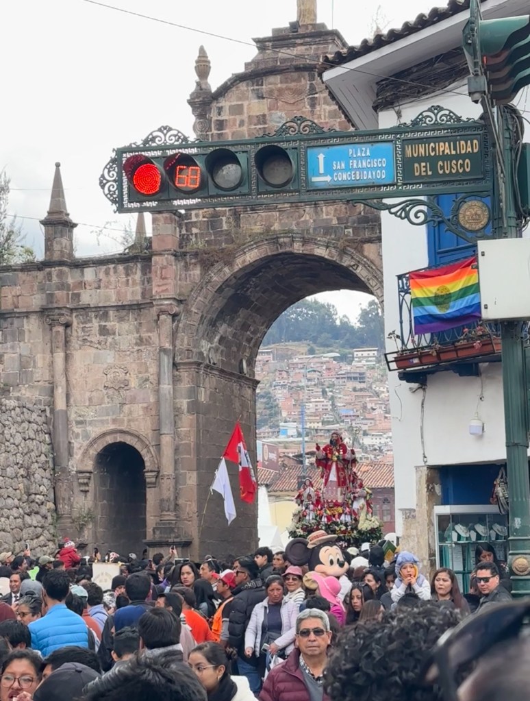 Busy street scene in Cusco, Peru, featuring an archway, traffic lights, and a vibrant crowd of people celebrating in the plaza.