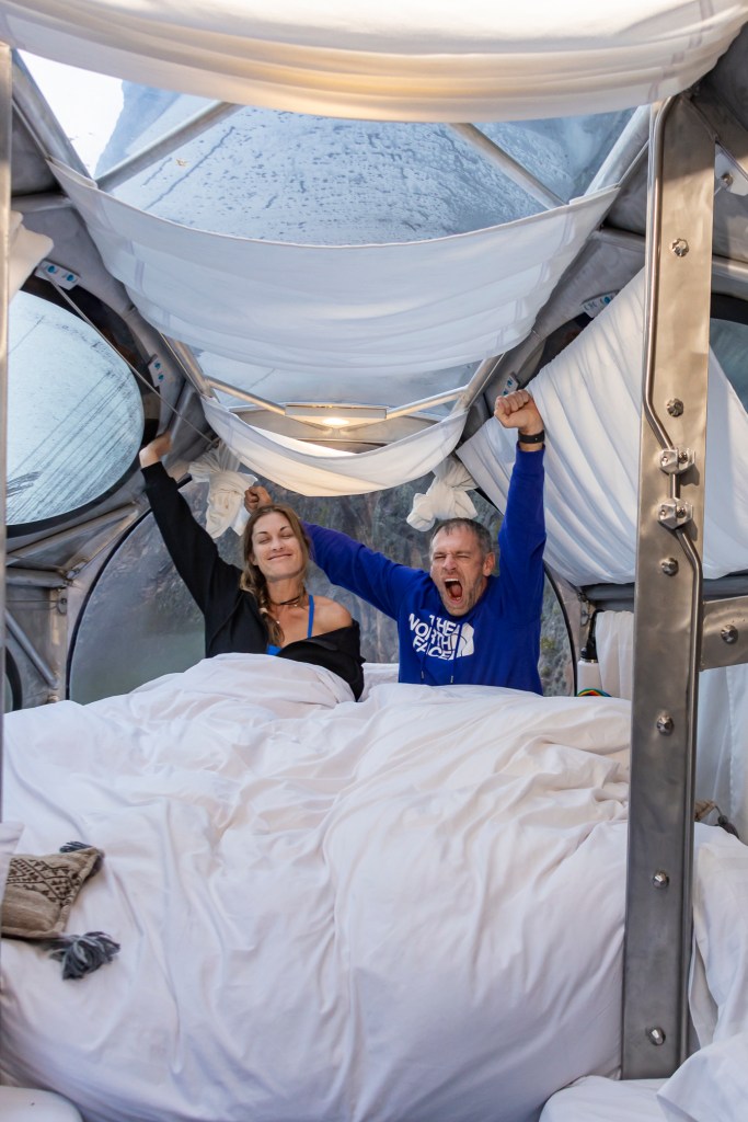 Two people in bed inside a transparent capsule hotel, joyfully stretching their arms in the air, surrounded by white bedding and a scenic view through the windows.