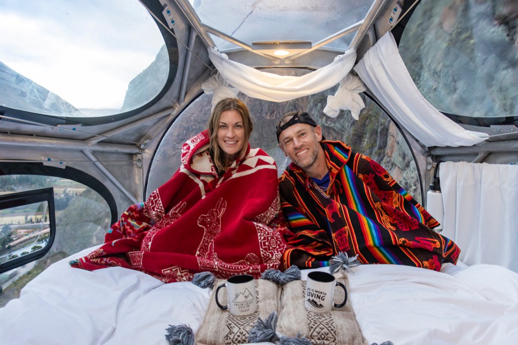 A couple sits inside a cozy capsule hotel high in the Sacred Valley, Peru. They are wrapped in colorful blankets, enjoying warm drinks, with scenic mountain views visible through large windows.