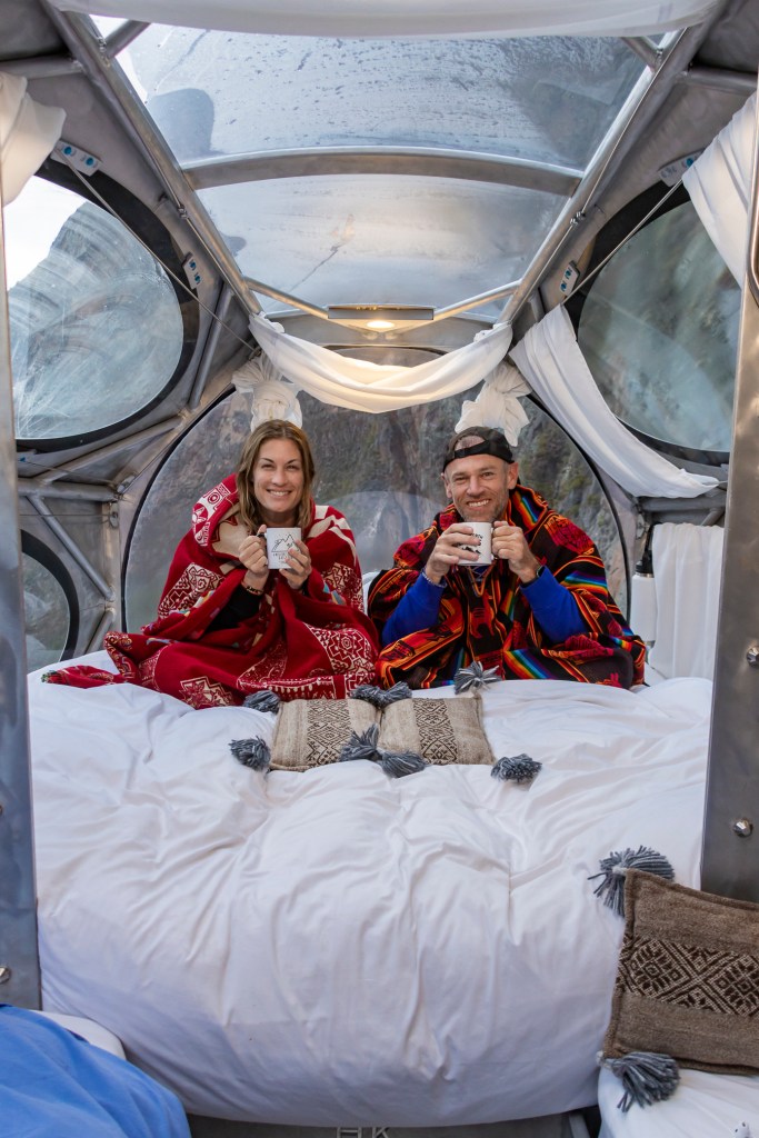 A couple sitting on a cozy bed inside a transparent capsule hotel in the Sacred Valley of Peru, wrapped in colorful blankets and holding warm mugs.