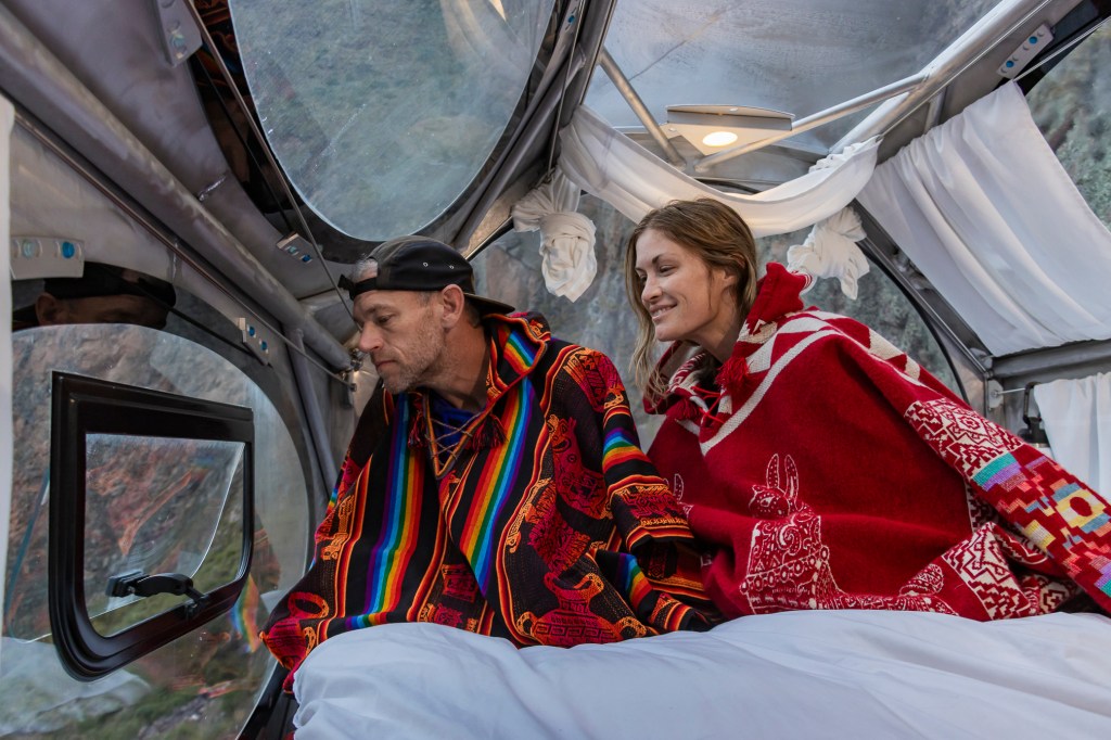 A couple in colorful traditional ponchos sitting in a glass capsule overlooking the Sacred Valley in Peru, with mountains visible through the transparent walls.