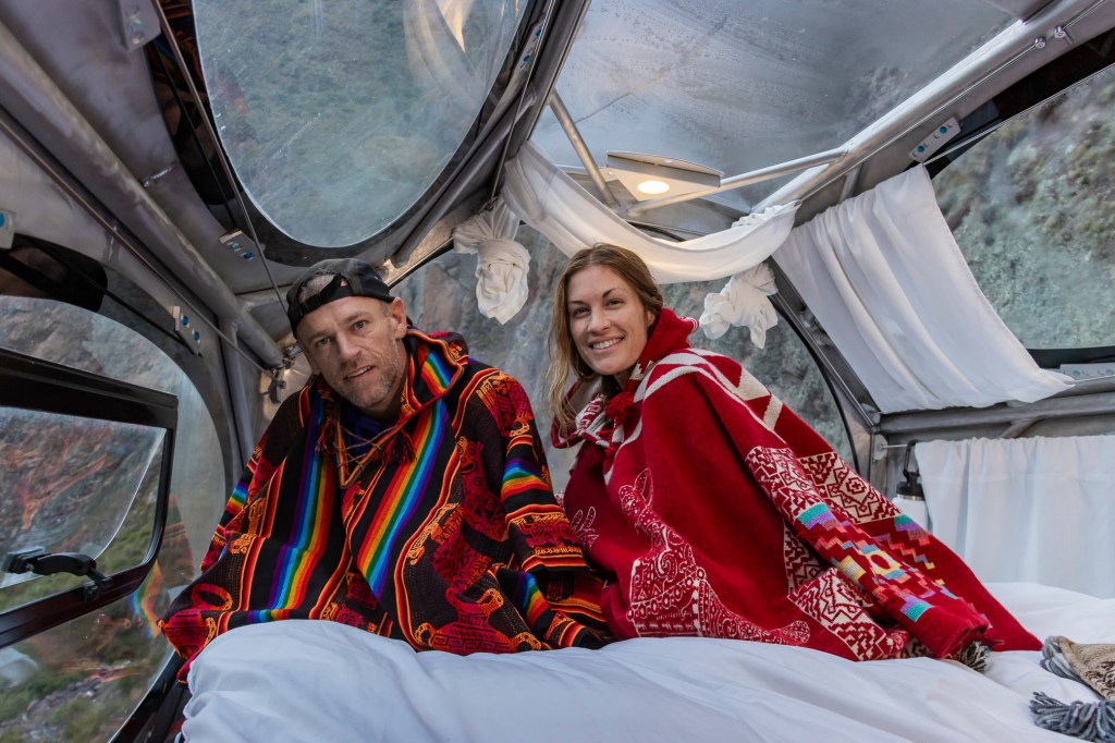 A couple sitting in a capsule hotel in the Sacred Valley, Peru, wearing colorful ponchos and smiling at the camera.